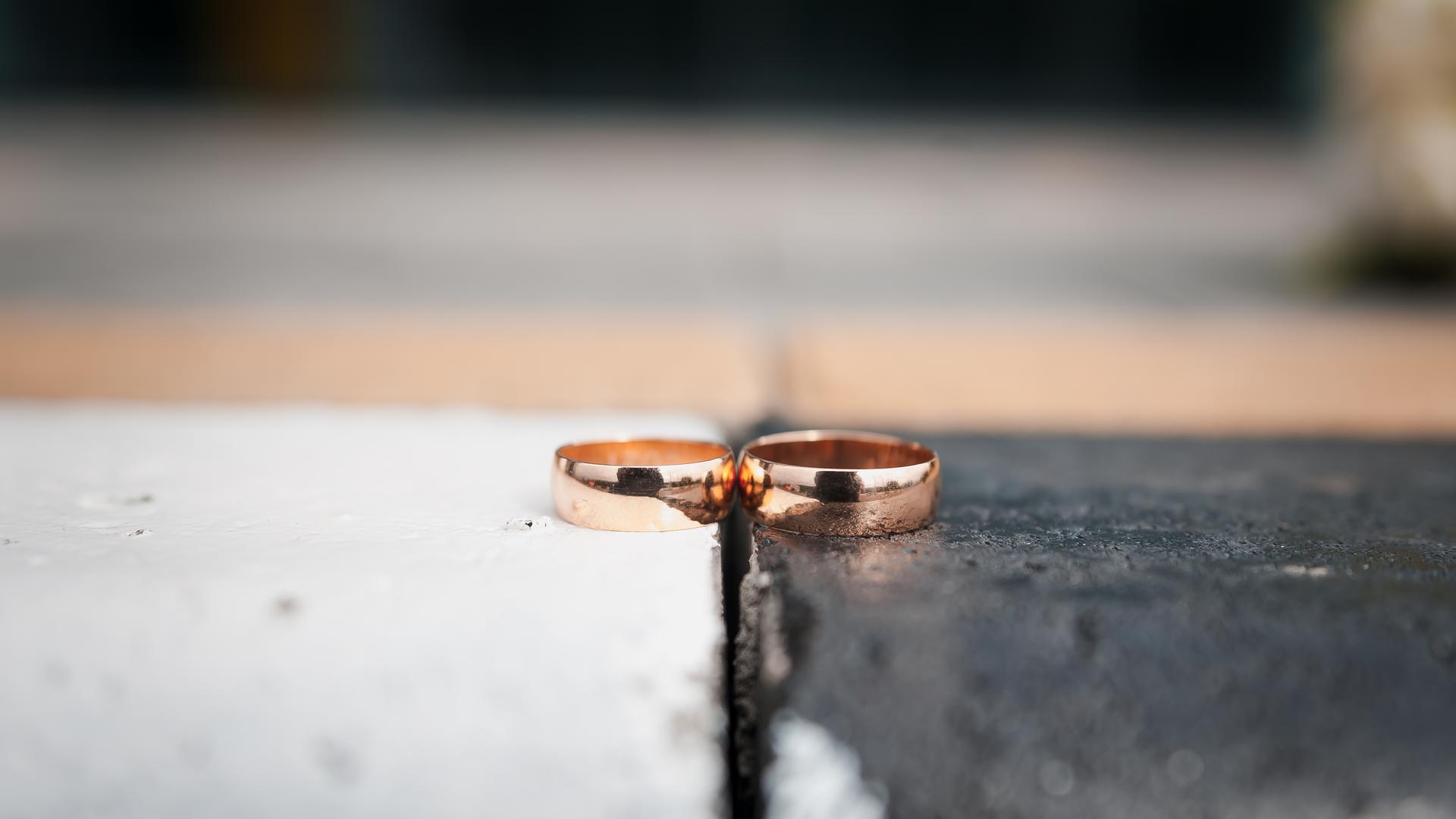 Zwei goldene Eheringe liegen nebeneinander. Der linke Ring liegt auf einem weißen, der rechte Ring auf einem schwarzen Untergrund.