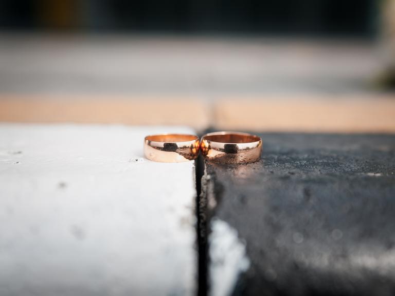 Zwei goldene Eheringe liegen nebeneinander. Der linke Ring liegt auf einem weißen, der rechte Ring auf einem schwarzen Untergrund.