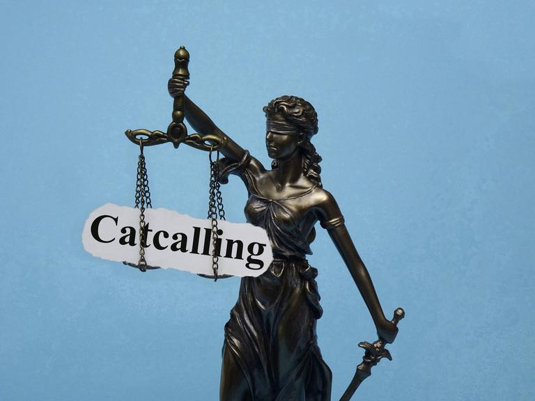 Eine Justizia-Stratue mit verbundenen Augen hat das Wort "Catcalling" auf den Waagschalen.