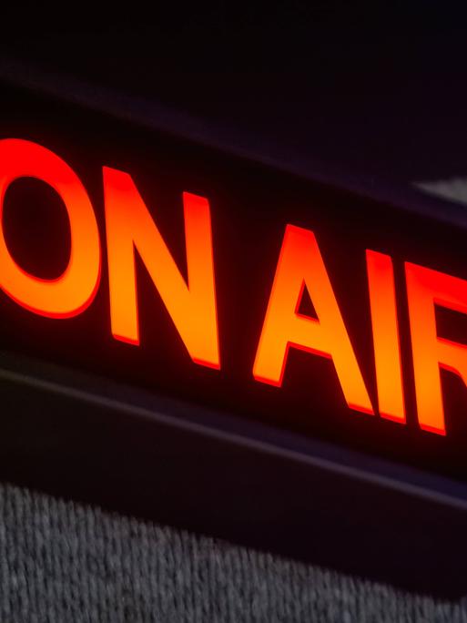 Ein rechteckiges ON AIR-Schild beleuchtet das Aufnahmestudio. Der leuchtend rote Schriftzug hebt sich von dem schwarzen Hintergrund ab. 