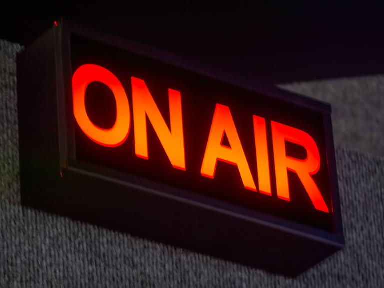 Ein rechteckiges ON AIR-Schild beleuchtet das Aufnahmestudio. Der leuchtend rote Schriftzug hebt sich von dem schwarzen Hintergrund ab. 