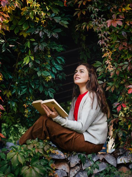 Porträt einer Frau, die lesend auf einer Mauer sitzt, die von wildem Wein umschlossen wird.