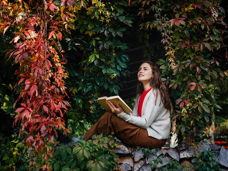 Porträt einer Frau, die lesend auf einer Mauer sitzt, die von wildem Wein umschlossen wird.
