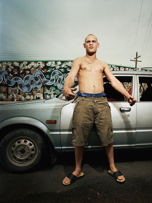 Ein junger Mann mit nacktem Oberkörper und grimmiger Mimik steht vor einem Auto.
