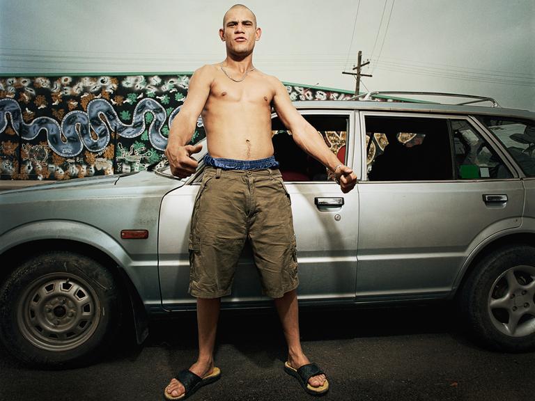 Ein junger Mann mit nacktem Oberkörper und grimmiger Mimik steht vor einem Auto.