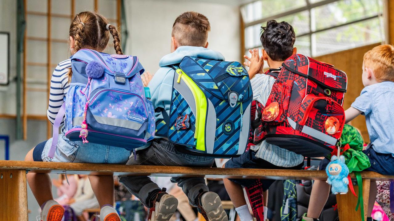 Zu sehen sind mehrere Schulkinder von hinten mit Ranzen, die bei einer Feier in einer Turnhalle sitzen.