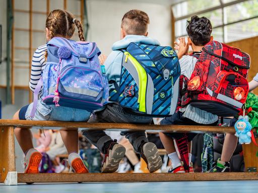 Zu sehen sind mehrere Schulkinder von hinten mit Ranzen, die bei einer Einschulungsfeier in einer Turnhalle sitzen.