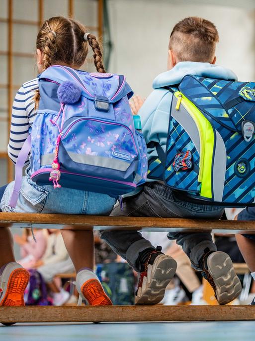 Zu sehen sind mehrere Schulkinder von hinten mit Ranzen, die bei einer Einschulungsfeier in einer Turnhalle sitzen.