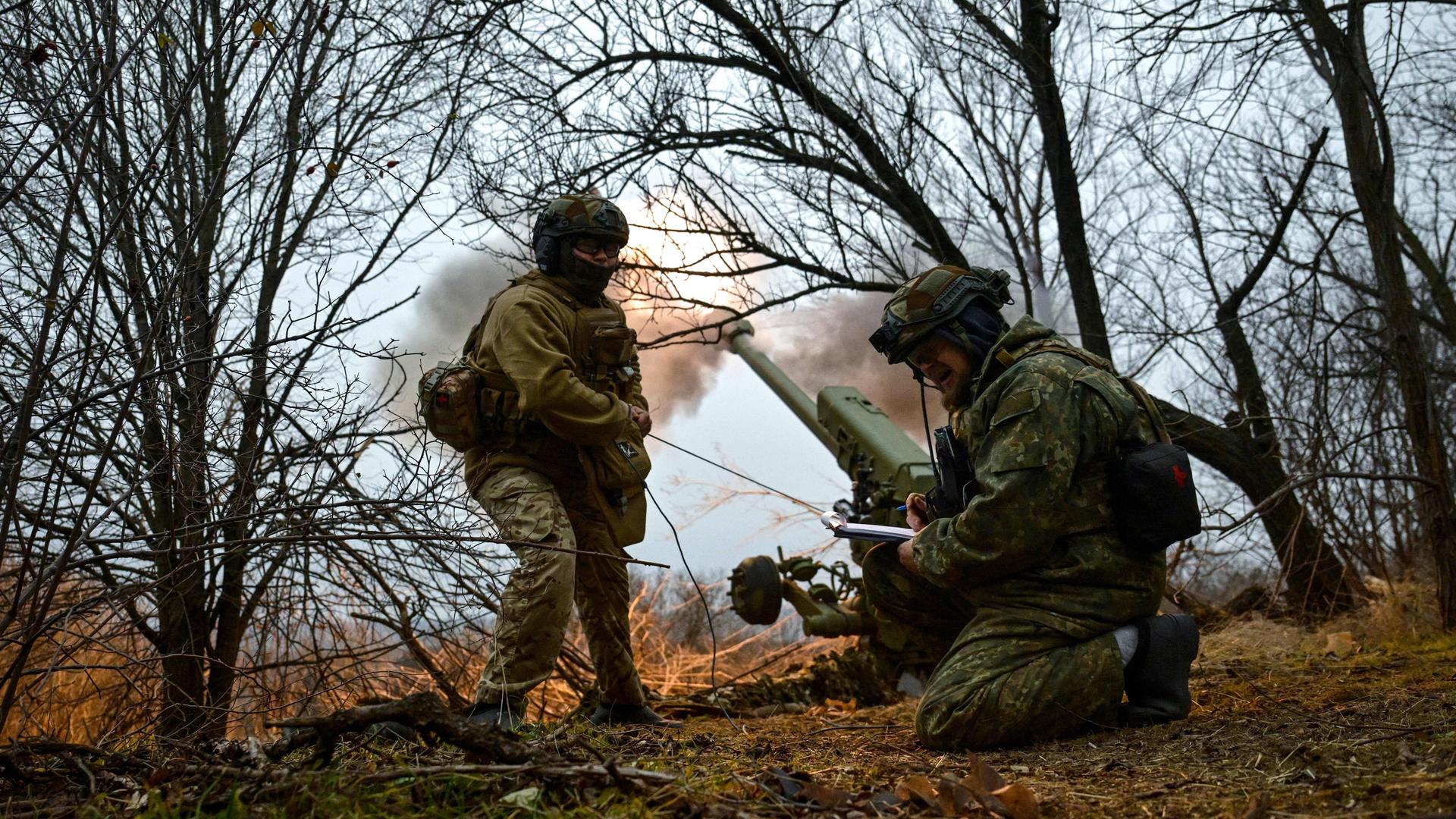 Ukrainische Soldaten im Flecktarn-Uniformen ducken sich in einem Wäldchen. Hinter ihnen feuert eine Haubitze eine Granate in Richtung der russischen Truppen. Der Rauch der Treibladung ist an der Mündung des Kanonenrohrs zu sehen. 