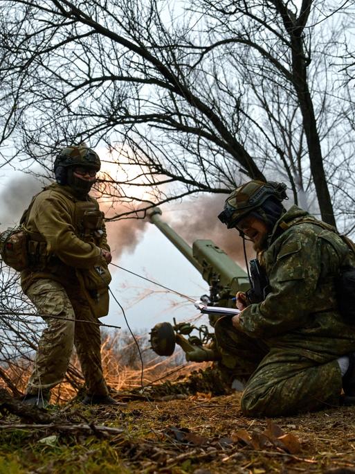 Ukrainische Soldaten im Flecktarn-Uniformen ducken sich in einem Wäldchen. Hinter ihnen feuert eine Haubitze eine Granate in Richtung der russischen Truppen. Der Rauch der Treibladung ist an der Mündung des Kanonenrohrs zu sehen. 