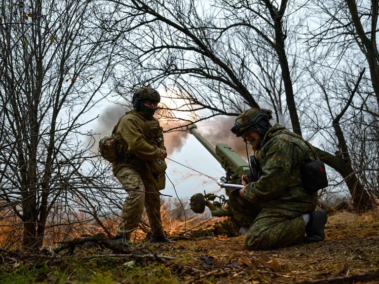 Ukrainische Soldaten im Flecktarn-Uniformen ducken sich in einem Wäldchen. Hinter ihnen feuert eine Haubitze eine Granate in Richtung der russischen Truppen. Der Rauch der Treibladung ist an der Mündung des Kanonenrohrs zu sehen. 