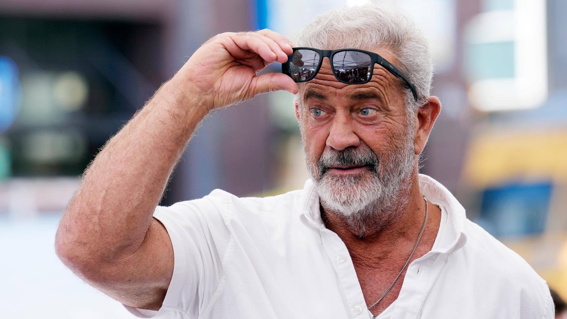 Mel Gibson - ein grauhaariger Mann mit Bart und weißem Poloshirt - hebt mit einer Hand seine Sonnenbrille zur Stirn und schaut überrascht. 