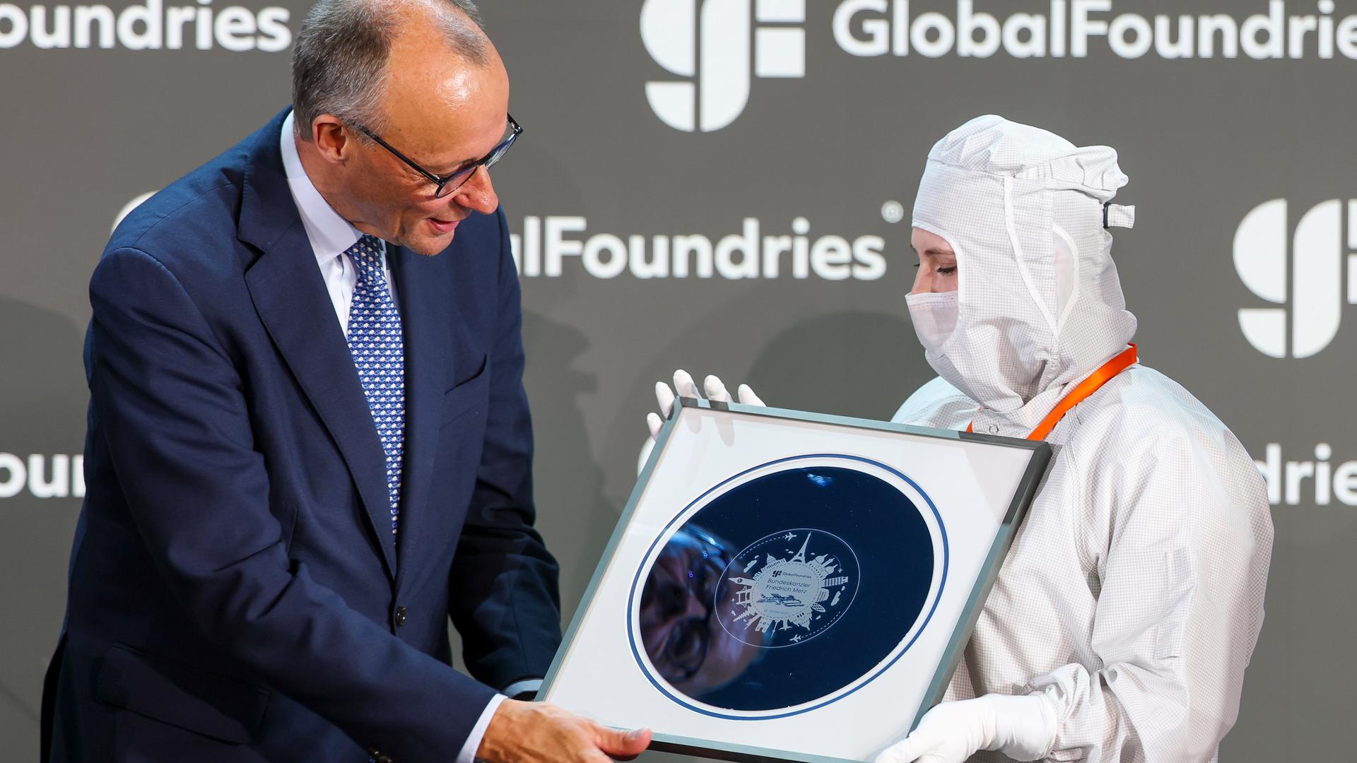 Eine junge Frau hat einen weißen Schutzanzug gegen Staub an. Sie überreicht Bundeskanzler Friedrich Merz eine Waferscheibe.
       Eine junge Frau hat einen weißen Schutzanzug gegen Staub an. Sie überreicht Bundeskanzler Friedrich Merz eine Waferscheibe.