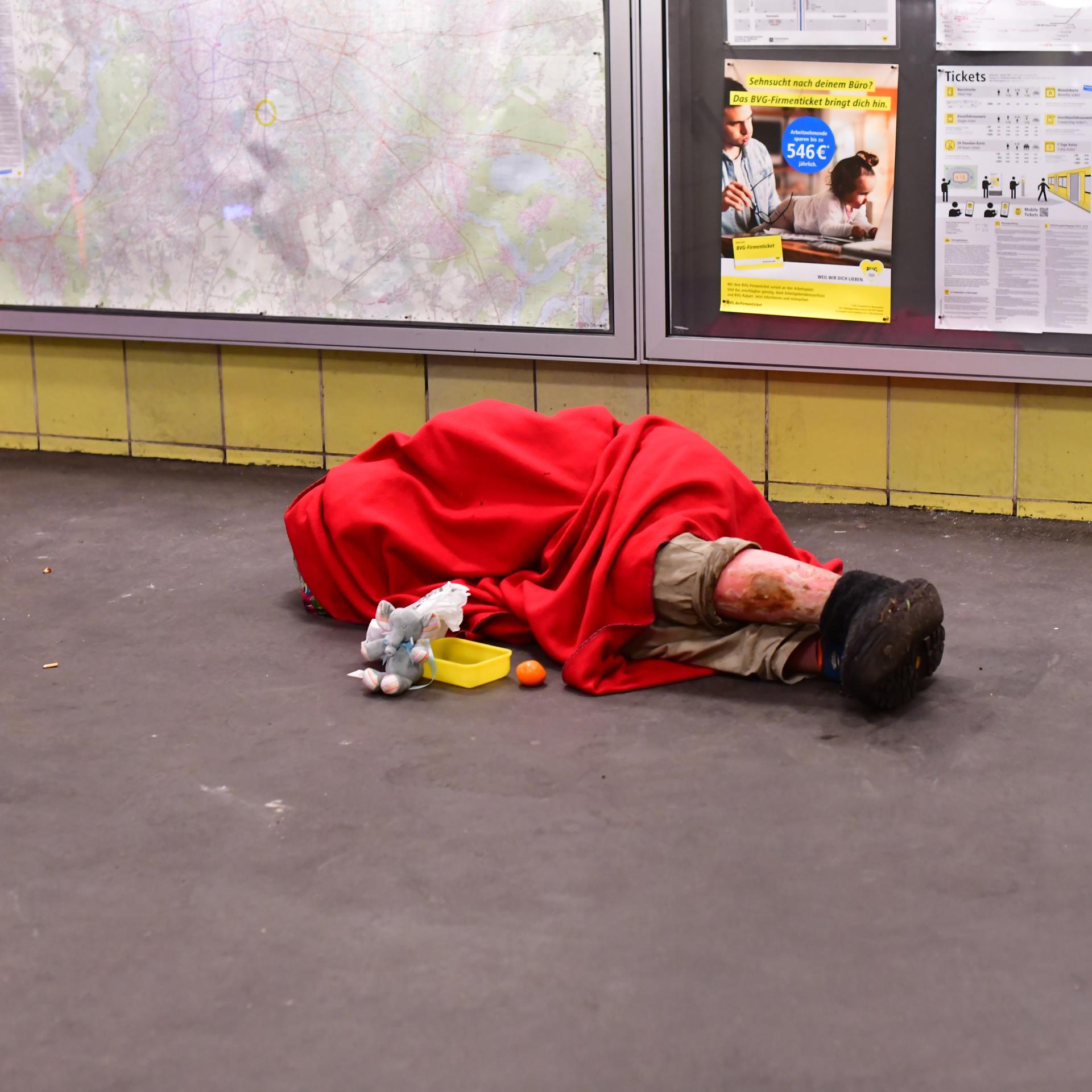 Ein Mensch ohne Obdach liegt in einen Schlafsack gehüllt morgens im Bahnhof Tempelhof mit einem Becher für Spenden vor sich und schläft auf dem Boden.