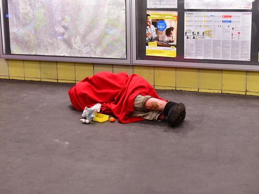 Ein Mensch ohne Obdach liegt in einen Schlafsack gehüllt morgens im Bahnhof Tempelhof mit einem Becher für Spenden vor sich und schläft auf dem Boden.