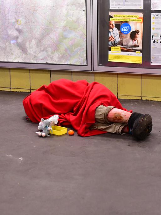 Ein Mensch ohne Obdach liegt in einen Schlafsack gehüllt morgens im Bahnhof Tempelhof mit einem Becher für Spenden vor sich und schläft auf dem Boden.