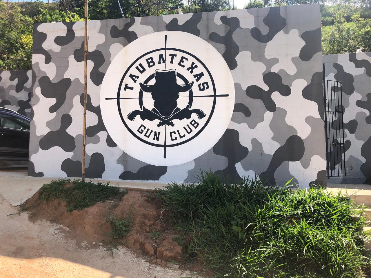 An einer Wand in grauen Tarnfarben prangt das Logo des Taubatexas Gun Club: Die schwarze Silhouette eines Mannes mit Hut im Fadenkreuz.