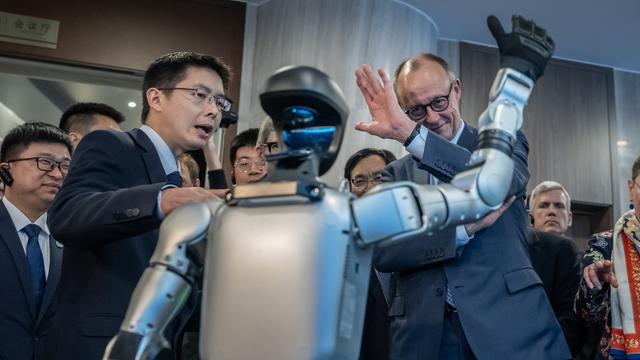 Bundes-Kanzler Merz steht vor einem Roboter. Merz und der Roboter erheben eine Hand. Es sieht aus als würden sich die beiden grüßen.