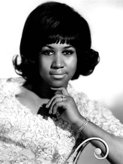 Aretha Franklin, die Queen of Soul, mit einem weißen Kleid auf einem Stuhl sitzend. Sie schaut in die Kamera.