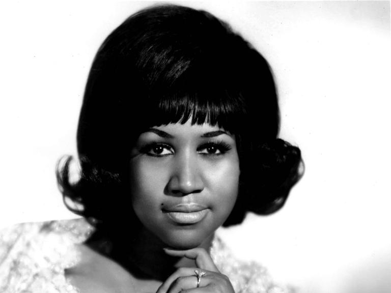 Aretha Franklin, die Queen of Soul, mit einem weißen Kleid auf einem Stuhl sitzend. Sie schaut in die Kamera.