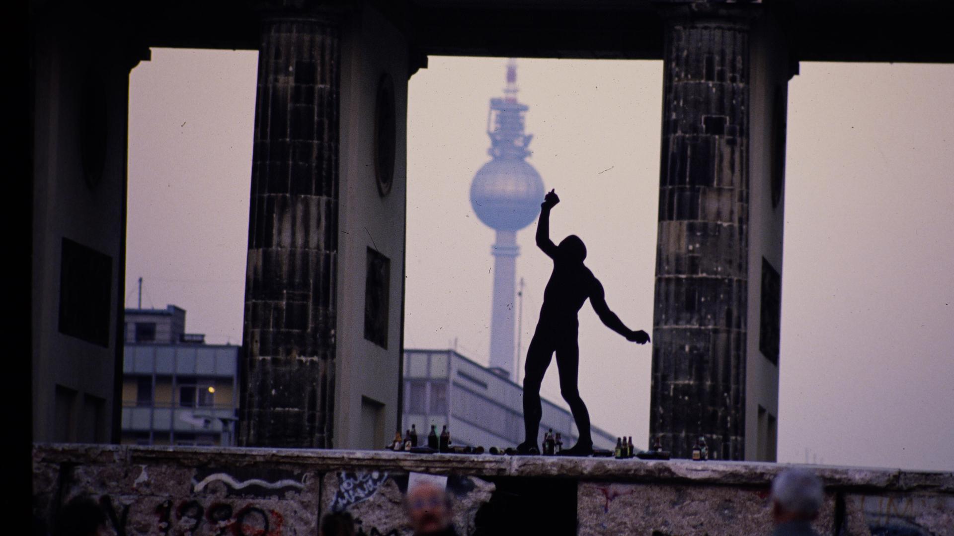 Ein Mann steht als Silhouette auf der Berliner Mauer zwischen den Säulen des Brandenburger Tors. Im Hintergrund ist der Berliner Fernsehturm zu sehen.