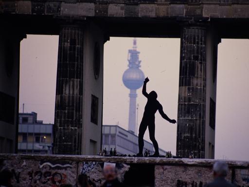 Ein Mann steht als Silhouette auf der Berliner Mauer zwischen den Säulen des Brandenburger Tors. Im Hintergrund ist der Berliner Fernsehturm zu sehen. Ein Mann steht als Silhouette auf der Berliner Mauer zwischen den Säulen des Brandenburger Tors. Im Hintergrund ist der Berliner Fernsehturm zu sehen.