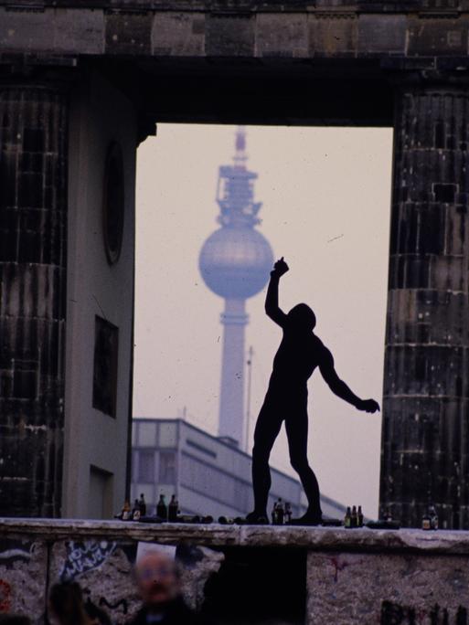 Ein Mann steht als Silhouette auf der Berliner Mauer zwischen den Säulen des Brandenburger Tors. Im Hintergrund ist der Berliner Fernsehturm zu sehen.