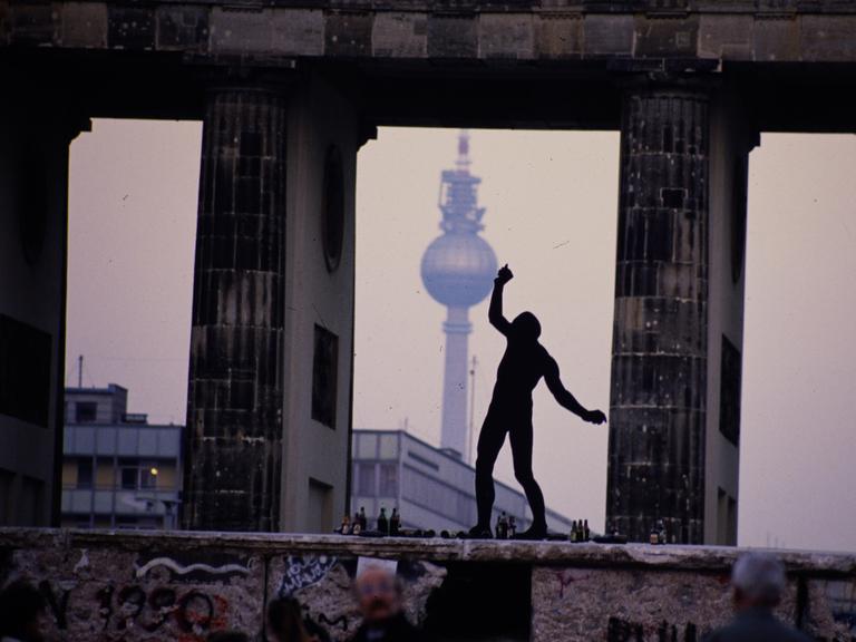 Ein Mann steht als Silhouette auf der Berliner Mauer zwischen den Säulen des Brandenburger Tors. Im Hintergrund ist der Berliner Fernsehturm zu sehen.