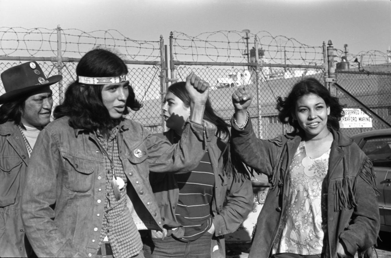 Eine Gruppe von jungen Native Americans steht vor dem Zaun von Alcatraz. Sie heben ihre Fäuste und schauen in die Kamera.