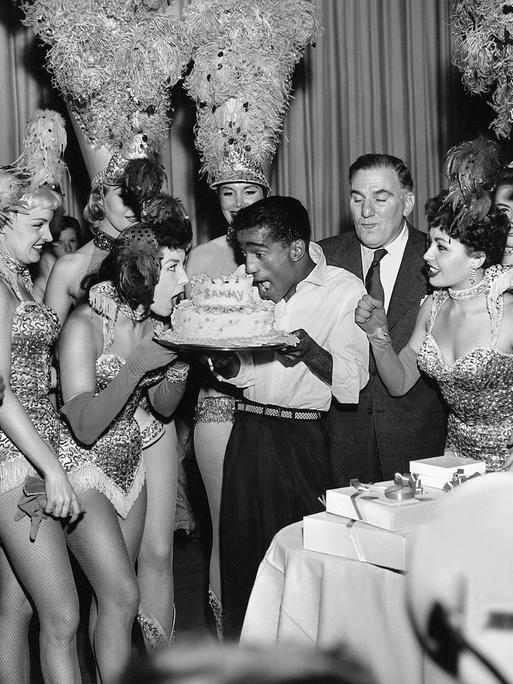 Sammy Davis Jr. & glitzernde Showgirls bei einer Pemiere in Las Vegas mit einer Torte auf der "Sammy" steht, 1955.