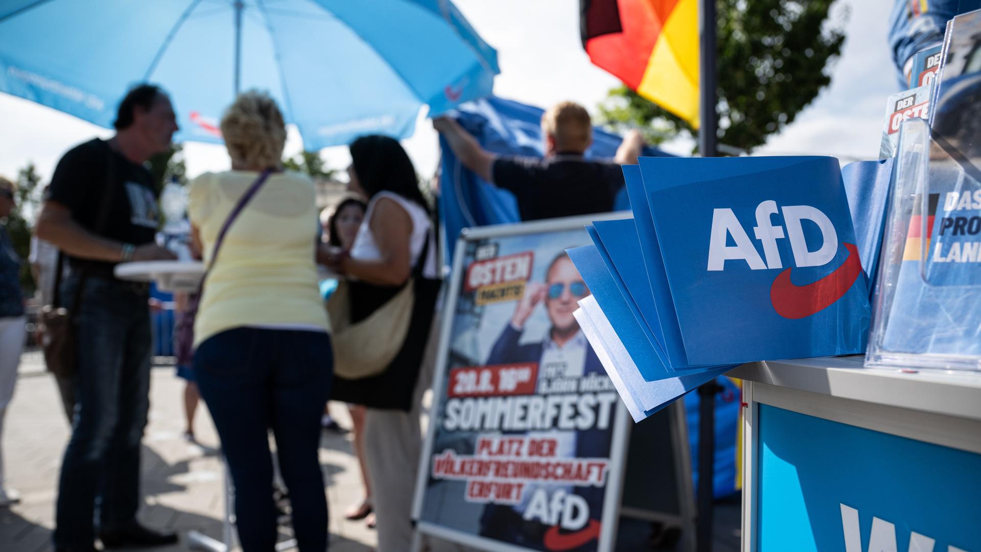 AfD-Wahlmaterial liegt an einem Stand aus. Verschwommen im Hintergrund: Menschen stehen an einem Stehtisch unter einem blauen AfD-Sonnenschirm und sprechen miteinander. Auch eine Deutschlandfahne ist zu sehen. 