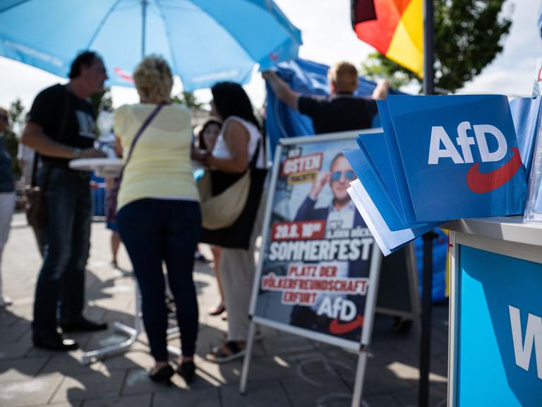 AfD-Wahlmaterial liegt an einem Stand aus. Verschwommen im Hintergrund: Menschen stehen an einem Stehtisch unter einem blauen AfD-Sonnenschirm und sprechen miteinander. Auch eine Deutschlandfahne ist zu sehen. 