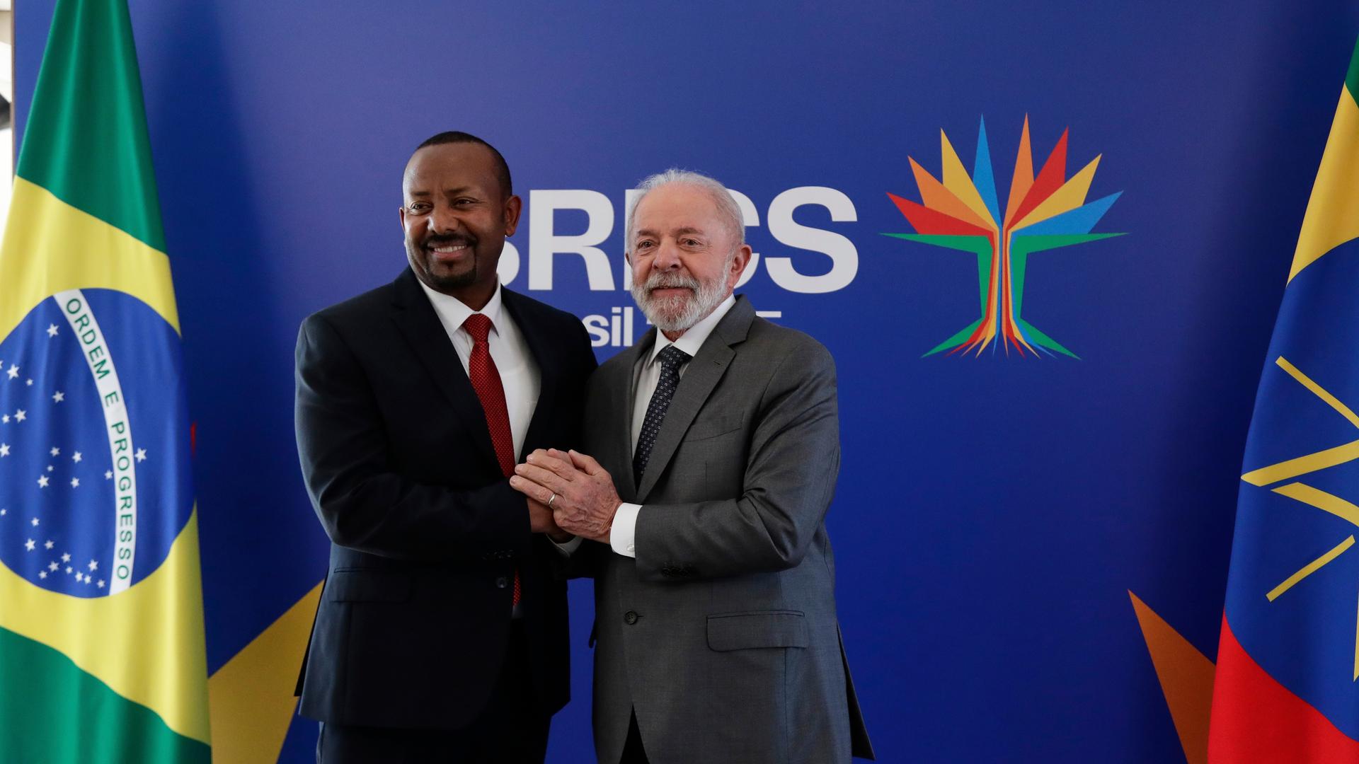 Brasiliens Präsident Lula da Silva und der äthiopische Premierminister Abiy Ahmed Ali schütteln die Hände und präsentieren sich den Kameras. Im Hintergrund ist das Logo der BRICS-Staaten zu sehen.
