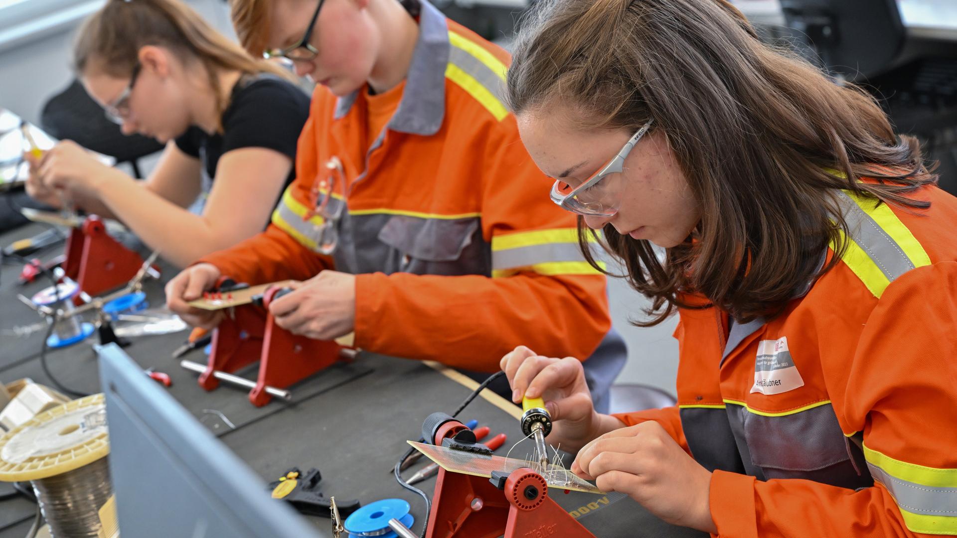 Auszubildende lernen im Elektrobereich vom Berufsbildungszentrum des Stahlproduzenten ArcelorMittal. Sie tragen orangfarbene Jacken und sitzen an einer Werkbank mit Lötkolben.