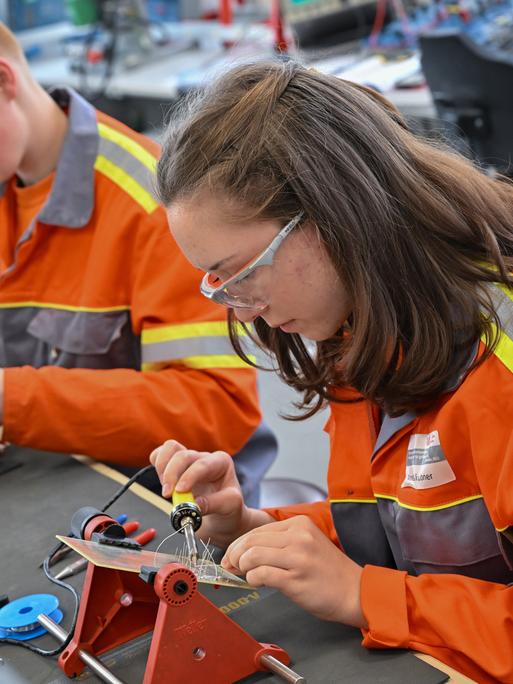 Auszubildende lernen im Elektrobereich vom Berufsbildungszentrum des Stahlproduzenten ArcelorMittal. Sie tragen orangfarbene Jacken und sitzen an einer Werkbank mit Lötkolben.