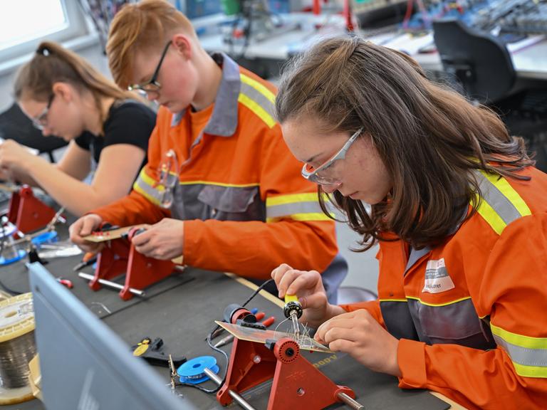 Auszubildende lernen im Elektrobereich vom Berufsbildungszentrum des Stahlproduzenten ArcelorMittal. Sie tragen orangfarbene Jacken und sitzen an einer Werkbank mit Lötkolben.