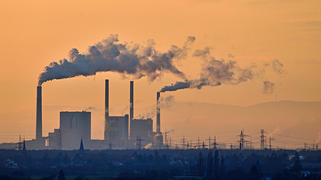 Mehrere Schornsteine einer Fabrik stoßen dichten grauen Rauch in einen orange‑goldenen Abendhimmel aus. Darunter liegen dunkle Gebäude, bläuliche Landschaft und Strommasten am Horizont. Mehrere Schornsteine einer Fabrik stoßen dichten grauen Rauch in einen orange‑goldenen Abendhimmel aus. Darunter liegen dunkle Gebäude, bläuliche Landschaft und Strommasten am Horizont.