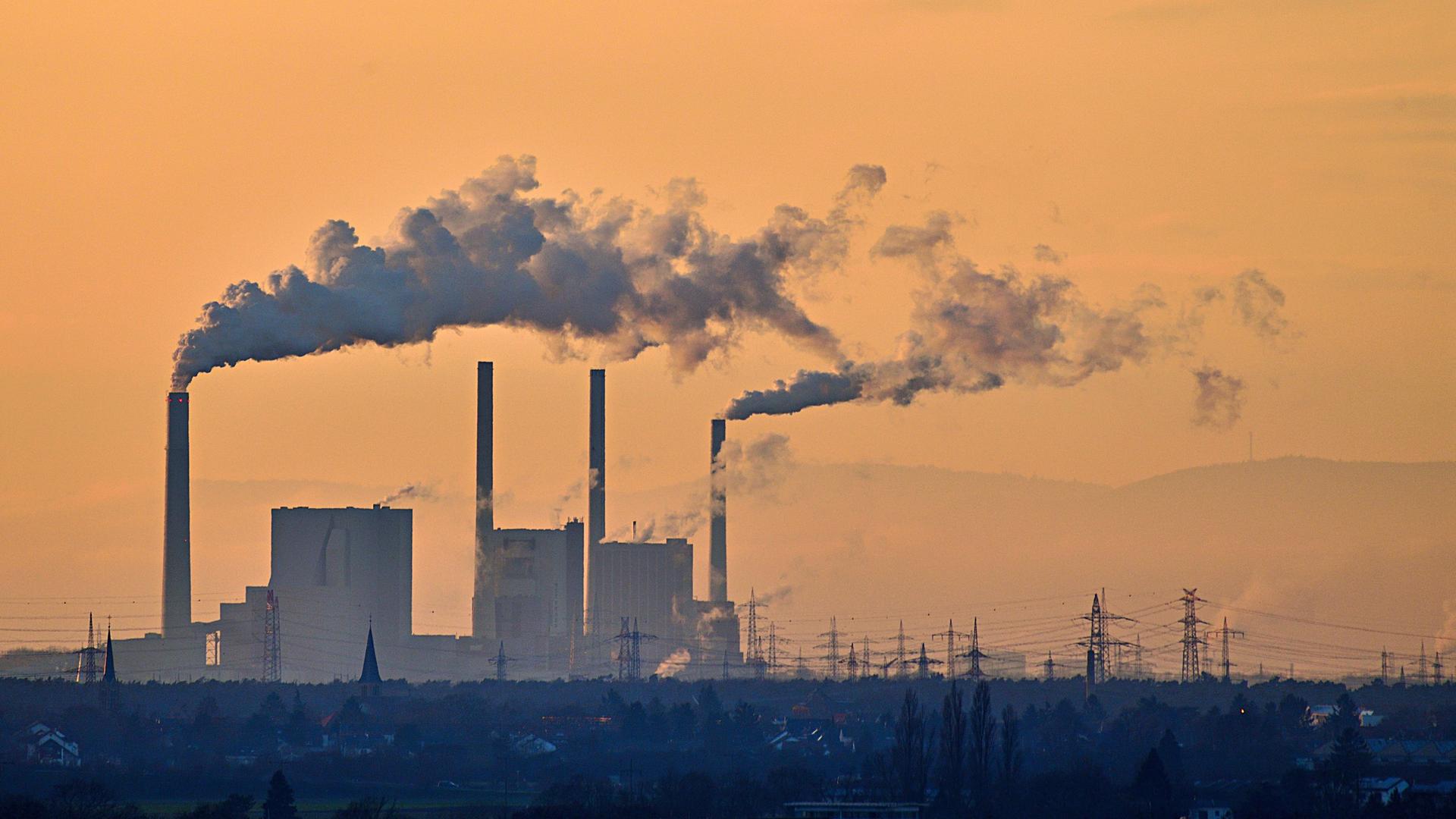 Mehrere Schornsteine einer Fabrik stoßen dichten grauen Rauch in einen orange‑goldenen Abendhimmel aus. Darunter liegen dunkle Gebäude, bläuliche Landschaft und Strommasten am Horizont.