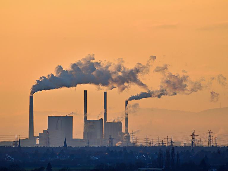 Mehrere Schornsteine einer Fabrik stoßen dichten grauen Rauch in einen orange‑goldenen Abendhimmel aus. Darunter liegen dunkle Gebäude, bläuliche Landschaft und Strommasten am Horizont.