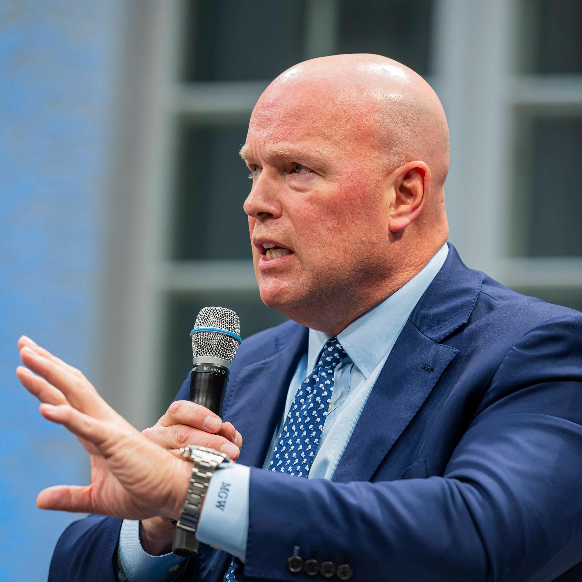 Matthew Whitaker, US-Botschafter bei der NATO, hat ein Mikrofon in rechten Hand und gestikuliert mit der linken, um seine Worte zu unterstreichen.
