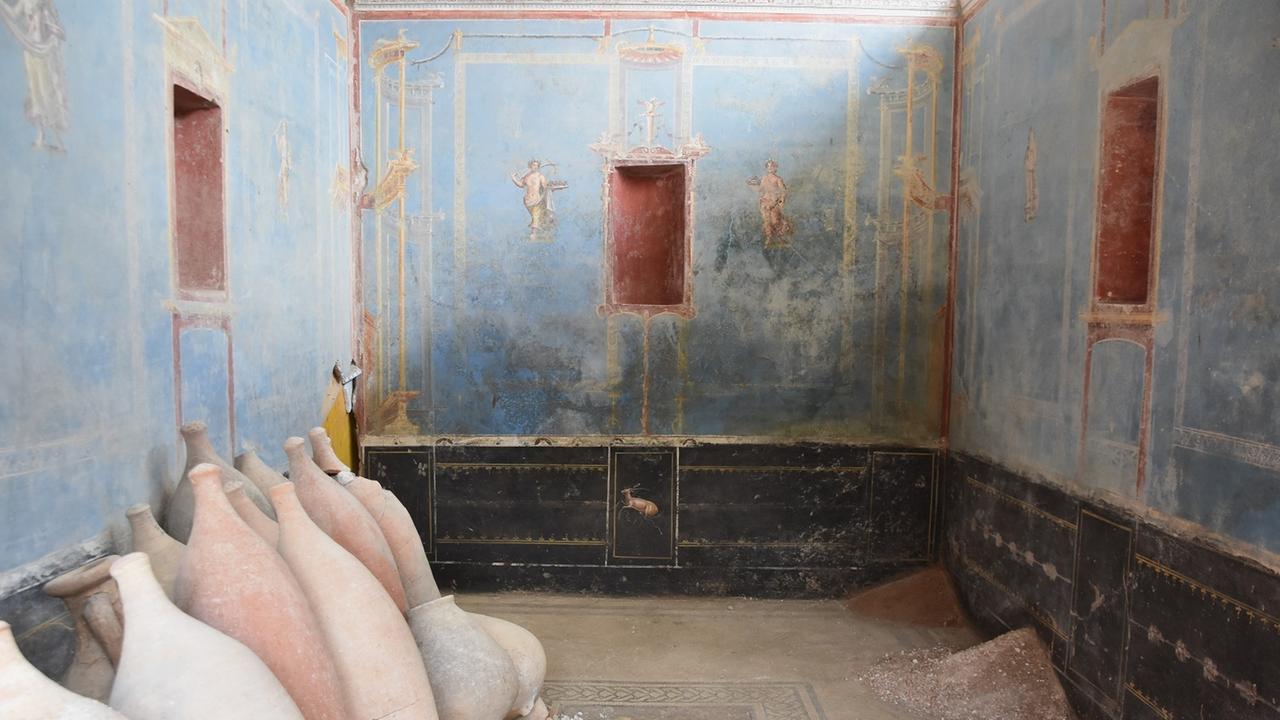 Ein Raum mit blauen Wänden und gemalten weiblichen Figuren ist in Pompeji zu sehen. Am Rand stehen Amphoren. Ein Raum mit blauen Wänden und gemalten weiblichen Figuren ist in Pompeji zu sehen. Am Rand stehen Amphoren.