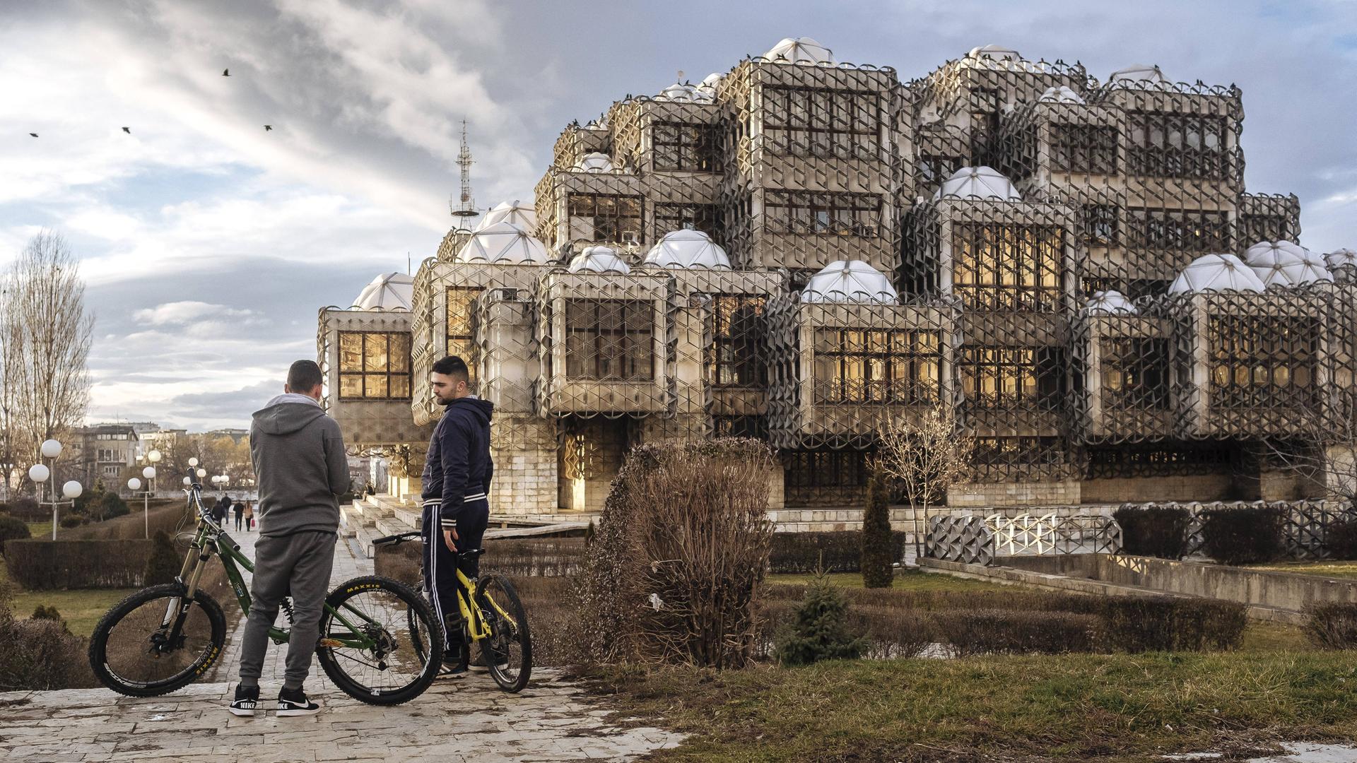 Zwei junge Männer stehen mit ihren Fahrrädern vor der Nationalbibliothek des Kosovo in Pristina.