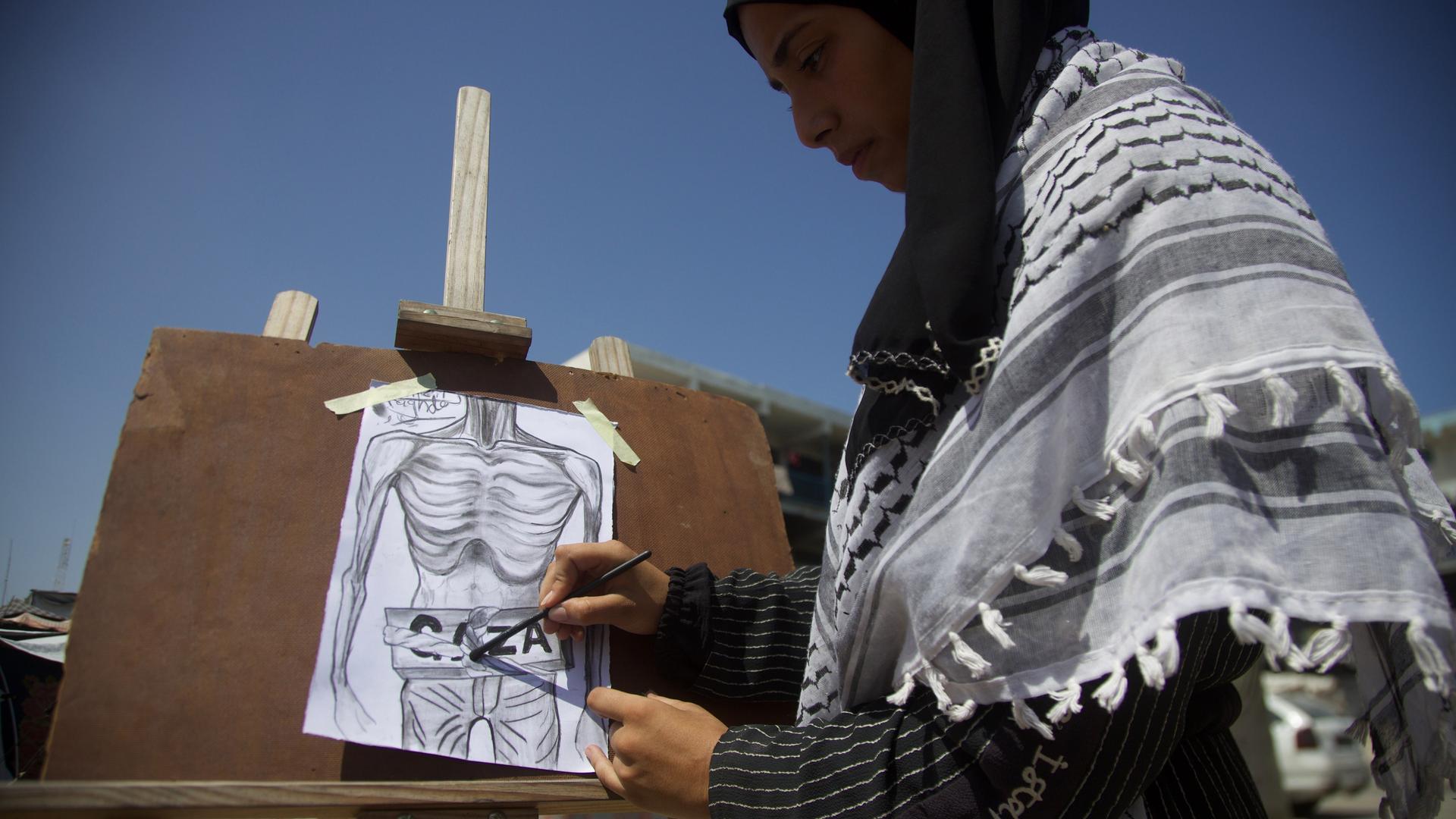 Eine Malerin steht an einer Staffelei. Ihr Bild zeigt einen bis auf das Skelett abgemagerten Oberkörper mit der Aufschrift "Gaza".