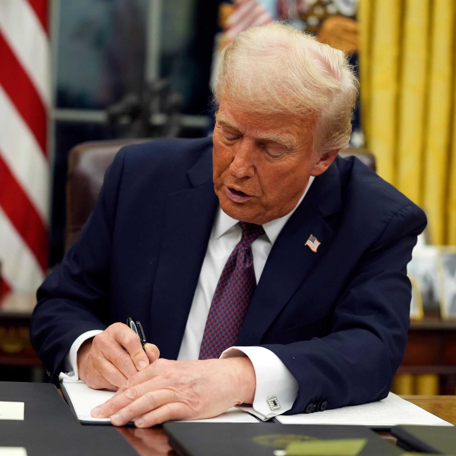 Donald Trump sitzt an einem Schreibtisch vor einer US-Fahne im Weißen Haus und unterzeichnet ein Dekret.