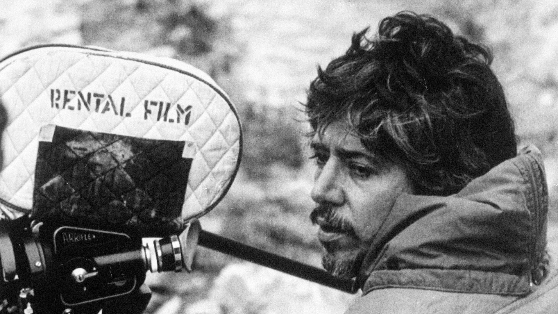 Der argentinische Filmemacher Luis Puenzo steht auf einem schwarz-weiß Foto neben einer Filmkamera.