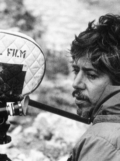 Der argentinische Filmemacher Luis Puenzo steht auf einem schwarz-weiß Foto neben einer Filmkamera.