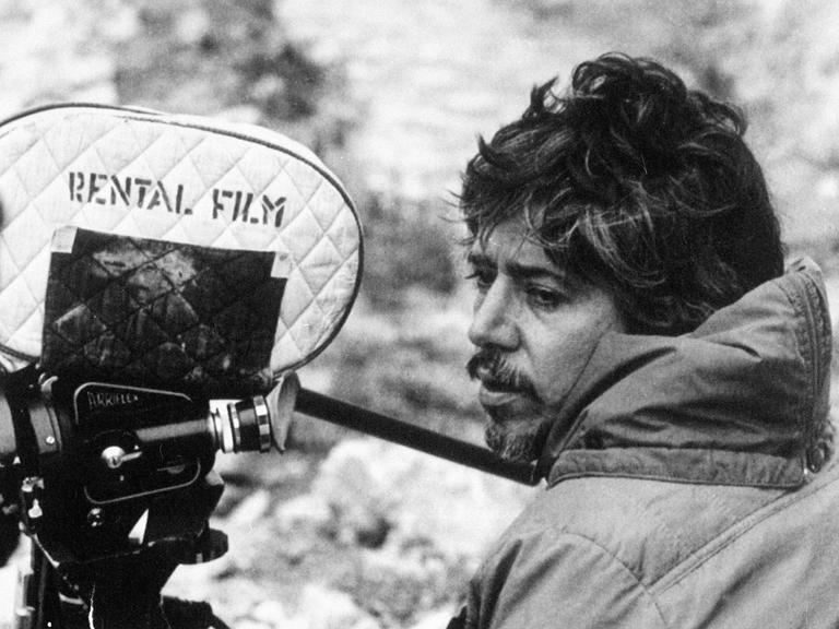 Der argentinische Filmemacher Luis Puenzo steht auf einem schwarz-weiß Foto neben einer Filmkamera.