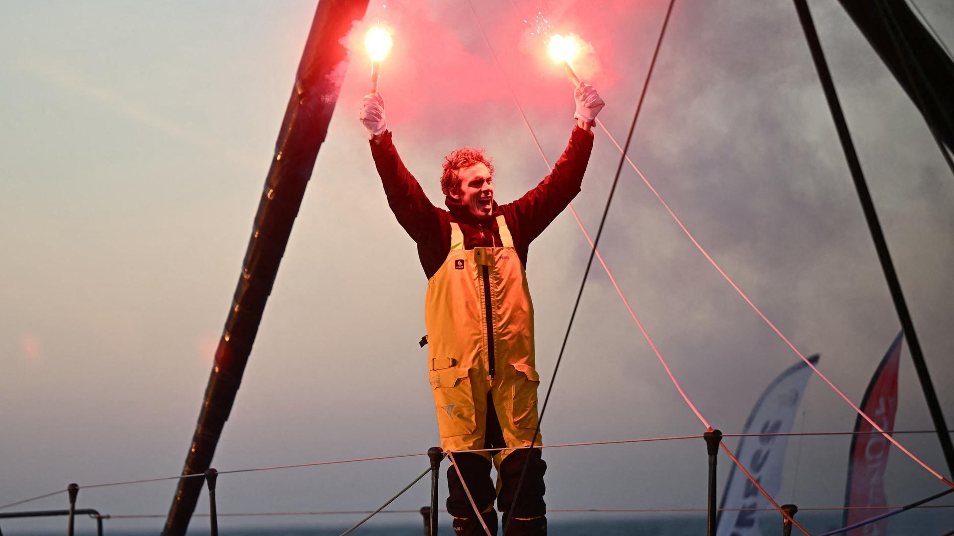  Charlie Dalin steht auf seinem Segel-Boot und hält zwei Leucht-Fackeln in die Luft.