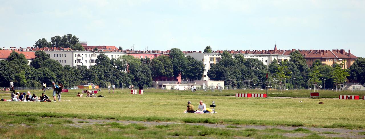 Menschen sitzen und stehen auf dem ehemaligen Flughafen Berlin-Tempelhof, nahe der Oderstraße im Schillerkiez in Neukölln, auf einer Wiese.