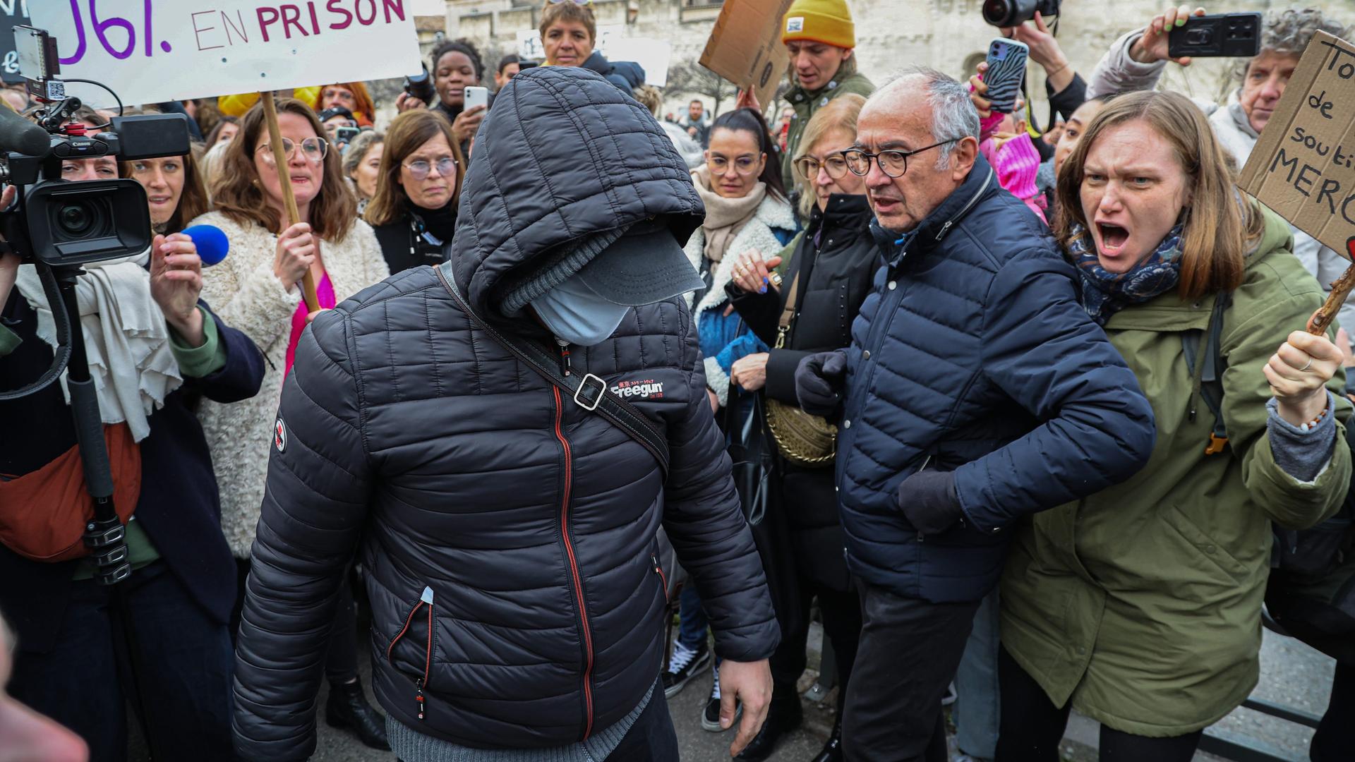 Ein maskierter Angeklagter im Vergewaltigungsprozess von Avignon geht zum Gericht. Um ihn herum sind wütende Menschen zu sehen.